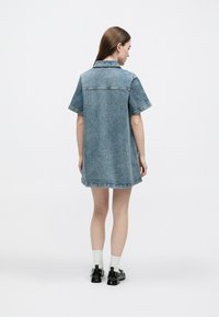 Φορέματα πουκαμίσου από τζιν με κοντά μανίκια, γιακά και άνετη εφαρμογή, σε μεσαίο μπλε με ξεβαμμένη υφή και γυρισμένο στρίφωμα. Μαύρα παπούτσια και λευκές κάλτσες.