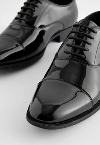 Schwarze Lackleder-Dressschuhe mit schlankem Design, Schnürverschluss und runder Zehenform, die über eine glatte Oberfläche und dezente Steppdetails verfügen.
