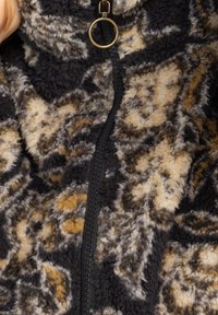 Chaqueta con cremallera de forro polar en negro, tostado y crema con un patrón floral, con cremallera negra y un anillo decorativo dorado. Textura suave.