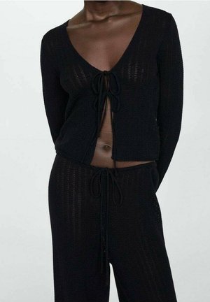 En person iført et sort strikket tohvids outfit med en dyb V-hals top bundet foran og matchende bukser med snoretræk.