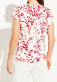 Kortærmet bluse i hvidt stof med et rødt blomstermønster. Bagudsidens visning viser tekstureret blondertrim på skuldrene og en rund bundkant.
