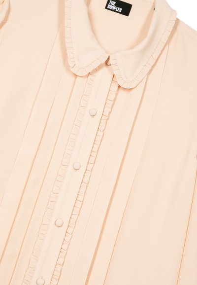Blusa crema con cuello redondeado, botones forrados de tela, pliegues verticales y ribete con volantes a lo largo de la tapeta de botones y los bordes del cuello.