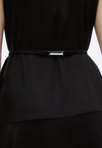 Μαύρη ζώνη από ύφασμα, με ορθογώνια μεταλλική αγκράφα. Ο σχεδιασμός είναι μινιμαλιστικός με καθαρή, λεία υφή.