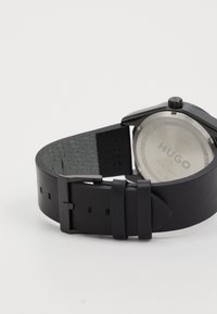 Μαύρη δερμάτινη λουρίδα ρολογιού με υφή στην εσωτερική επιφάνεια, μεταλλικό κούμπωμα και στρογγυλή θήκη. Στην πλάτη φαίνεται το όνομα μάρκας "HUGO" χαραγμένο σε ασημί.