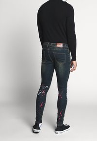 Vaqueros de mezclilla en azul oscuro con un ajuste ajustado, que presentan detalles de salpicaduras de pintura blanca y rosa en las piernas, y una etiqueta de cuero con la marca en la cinturilla.