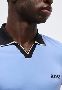 Κοντινό πλάνο ενός άνδρα που φοράει ανοιχτό μπλε πόλο μπλουζάκι με μαύρο γιακά και λογότυπο «BOSS» στο στήθος, δείχνοντας μέρος της γνάθου και του λαιμού του.