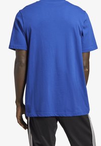 Camiseta de algodón azul de manga corta con un cuello redondo y una textura suave, vista desde atrás. Se pueden ver pantalones negros con rayas blancas.
