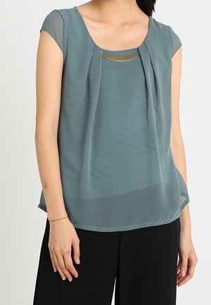 Kobieta w turkusowej, krótkim rękawem bluzce z szyfonu z plisami, nałożonej na dopasowany top oraz czarne szerokie spodnie.