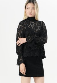 Črna čipkasta majica z visokim ovratnikom in razširjenimi rokavi, ki vsebuje cvetlične vzorce in valovit rob. Ujemanje s črno krilom.