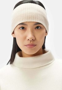 Beige ribbad pannband gjort av stickat material, bärs av en person med rakt svart hår. Mjuk textur med en tät passform runt pannan.