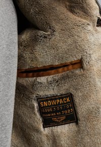 Nep bont voering in bruin met een gladde textuur; grijze buitenstof; heeft een label met de tekst "SNOWPACK," met zorginstructies, genaaid aan de binnenkant.