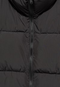 Gros plan d'une veste matelassée noire avec une fermeture éclair noire centrée qui court verticalement. Le tissu a une texture lisse et matelassée.