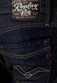 Vaqueros de mezclilla azul con un lavado oscuro, que presentan costuras prominentes, un parche de logo de cuero y un sutil acento rojo en el bolsillo trasero.