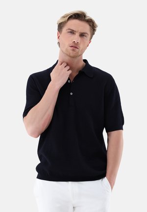 Junger Mann mit hellem Haar, trägt ein schwarzes kurzärmeliges Poloshirt und weiße Hose, eine Hand berührt den Hals, die andere in der Tasche.