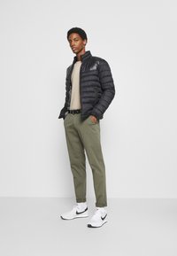 Zwarte pufferjack met een hoge kraag, gedragen over een beige trui, gecombineerd met olijfgroene broek en witte sneakers met zwarte accenten.