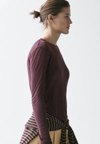 Femme aux cheveux châtain clair attachés en chignon, portant un haut à manches longues bordeaux, un pantalon beige et un pull rayé noué autour de la taille.