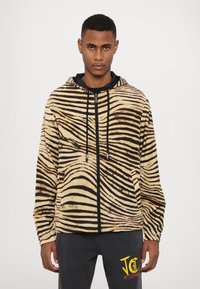Šviesiai rusvos ir juodos juostelės zip-up džemperis su lengvu audiniu, su raišteliais ir šoninėmis kišenėmis, vilkimas virš juodos marškinėlių.