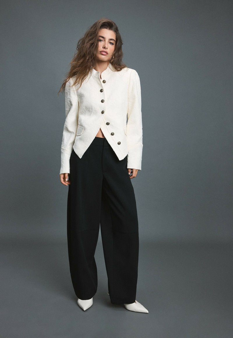 Jeune femme aux longs cheveux ondulés portant une veste blanche boutonnée, un pantalon large noir et des chaussures blanches pointues sur un fond gris.