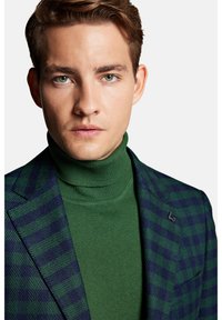 Zielona w kratę marynarka z niebieskimi akcentami, zestawiona z gładkim zielonym golfem. Posiada przypinkę na klapie i teksturowany materiał.