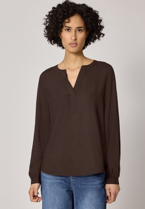 Kvinde med krøllet hår iført en mørkebrun bluse med lange ærmer og blå jeans, stående foran en ensfarvet, lysegrå baggrund.