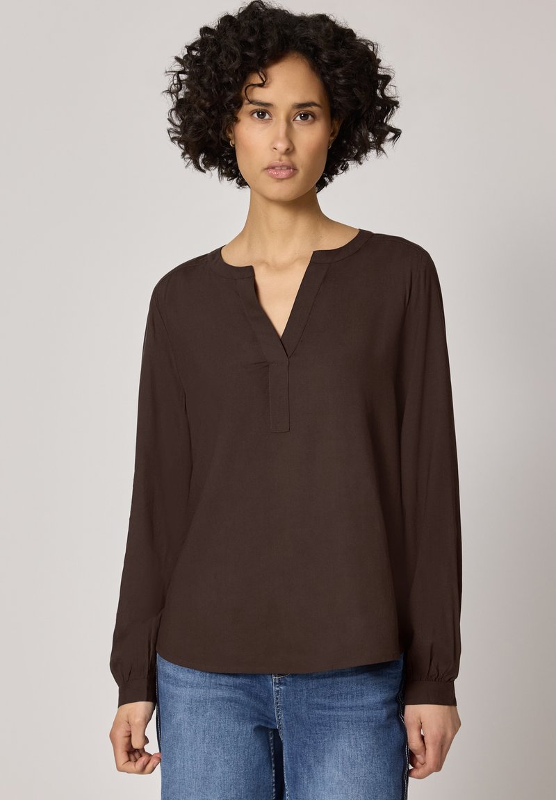 Femme aux cheveux bouclés portant un chemisier à manches longues marron foncé et un jean bleu, debout devant un fond uni gris clair.