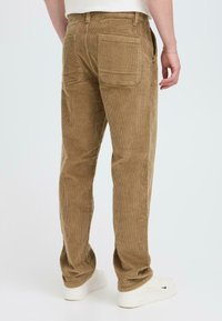 Corduroy pants in a warm beige color, featuring vertical ribbed texture, two back pockets, and slim silhouette. Worn with white sneakers.