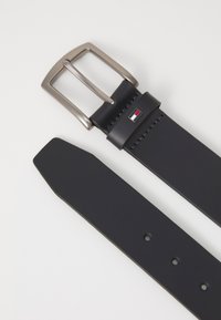 Ceinture en cuir bleu marine avec une boucle rectangulaire en argent, arborant un petit logo coloré et des coutures contrastées. Se termine par une forme effilée.