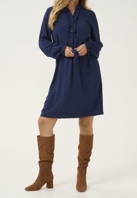 Abito blu navy con colletto arricciato, chiusura a bottoni e maniche lunghe. Abbinato a stivali alti in suede marrone con tacco a blocco.