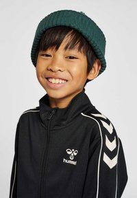 Chaqueta negra con cremallera y diseño de chevron blanco en las mangas, con logo en el pecho. Gorro de punto verde con textura acanalada; niño sonriente.