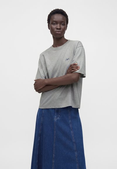 Femme aux cheveux courts portant un t-shirt gris à manches courtes et une longue jupe en jean bleue, debout les bras croisés devant un fond uni.