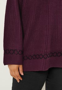 Pull en tricot violet avec une texture côtelée, présentant un motif en losanges noirs le long de l'ourlet et des poignets. Un bras visible reposant sur un pantalon noir.