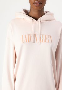 Svetlo roza kapuca iz mehke tkanine, z belim in breskev roza logotipom "CALVIN KLEIN JEANS" spredaj. Nastavljiv kapuco z vrvico.