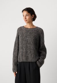 Przytulny, oversize'owy sweter w ciemnoszarym wełnianym kolorze z falistym wzorem, lekką teksturą i prążkowanymi mankietami. Połączony z szerokimi czarnymi spodniami.
