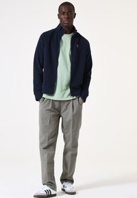 Marineblaue Zip-Jacke über einem hellgrünen T-Shirt, kombiniert mit grauen Hosen und weißen Sneakern mit schwarzen Streifen; das Modell steht in der Mitte.