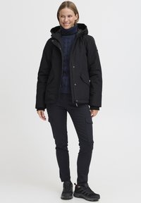 Veste isolante noire avec capuche, associée à un col roulé en tricot à câbles bleu marine, un pantalon cargo noir et des chaussures de sport noires.
