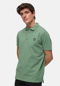 Grön piké skjorta i bomull, med en treknapplist, korta ärmar och ett litet marinblått emblem på vänster bröst.