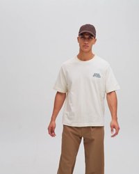 Lys beige bomulds t-shirt med et trykt logo på venstre bryst, parret med brune straight-leg bukser og en mørk kasket.