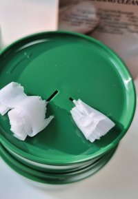 Zielony okrągły pojemnik z gładką pokrywką, na której wystają dwa białe paski woskowe z szczelin. Pojemnik ma błyszczące wykończenie.