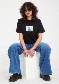 Femme aux cheveux bouclés portant des lunettes de soleil noires, un t-shirt noir graphique, un jean large bleu, plusieurs bagues et bracelets, assise sur un bloc blanc.