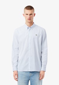 Camisa de manga larga a rayas en azul claro y blanco, con cuello abotonado y pequeño logo verde en el pecho. Tejido de algodón.