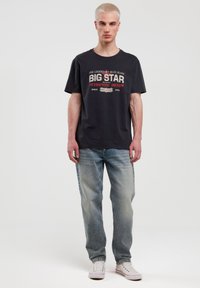Juodas medvilnės marškinėlis su spaudiniu "BIG STAR AUTHENTIC DENIM" logotipu, derinamas su šviesiai mėlynomis blukusiomis džinsomis ir baltais sportiniais bateliais.