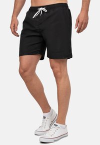 Pantaloni da bagno neri con vita elastica e lacci bianchi, con logo laterale e vestibilità comoda. Indossati con sneakers in tela bianche.