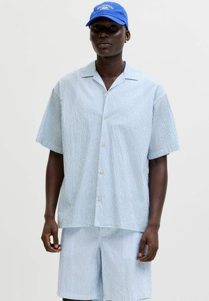 Temnopolta oseba nosi svetlo modro-belo črtasto srajco s kratkimi rokavi in gumbi ter ujemajoče kratke hlače s modro kapo.