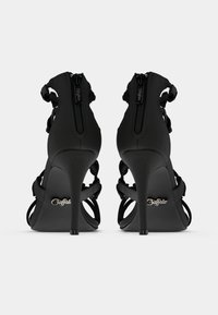 Melnas augstpapēžu sandales ar aizmugurējām rāvējslēdzējām un potīšu siksnām, skatītas no aizmugures uz vienkārša balta fona.