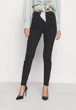 Vrouw draagt zwarte skinny jeans met hoge taille, zwarte puntige hoge hakken en een lichtgroene top met lange mouwen, gestrikt in de taille.