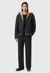 Schwarze Lederjacke mit beigem Lammfellfutter, Frontreißverschluss, zwei vordere Taschen, kombiniert mit schwarzen Hosen und Schuhen.