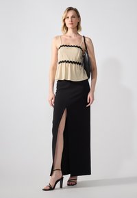 Beige peplum topp med svarte bølgete kanter, kombinert med et svart skjørt med høy splitt. Tilbehør inkluderer en svart håndveske og stroppehæler.