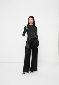 Top lungo a maniche lunghe con motivo nero abbinato a pantaloni larghi in satin neri; la modella tiene in mano una piccola borsa bianca e indossa sandali neri con cinturini.