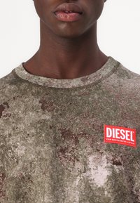 Nahaufnahme einer Person, die ein grün-braunes, strukturiertes Hemd mit einem rot-weißen Diesel-Logo-Patch auf der Brust trägt.