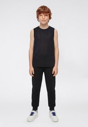 Roodharige jongen staand, gekleed in een zwart mouwloos mesh-shirt, zwarte joggingbroek en witte sneakers met blauwe veters tegen een effen achtergrond.